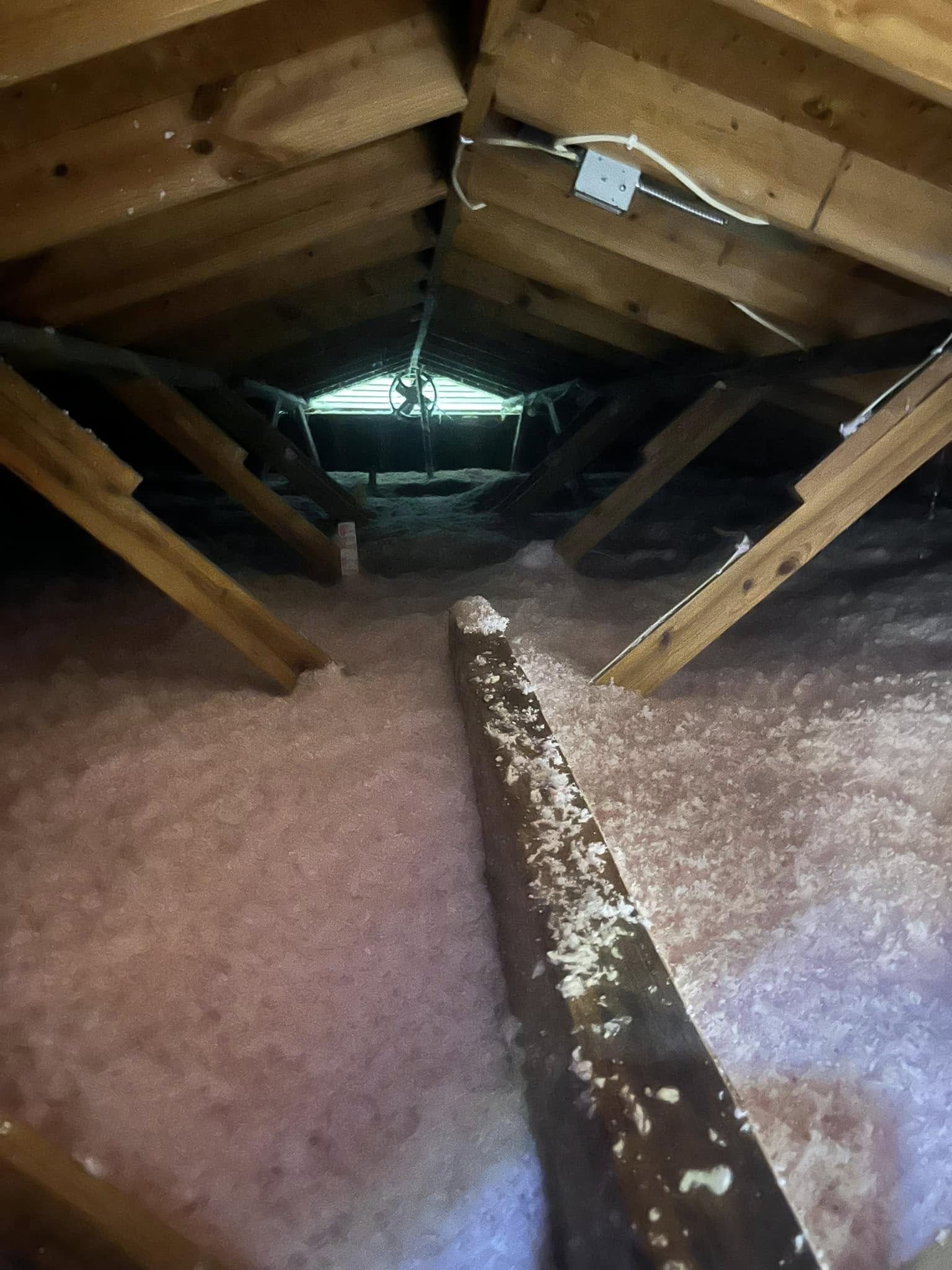 An attic with exposed wooden beams, pink insulation covering the floor, and a single light fixture on the ceiling.