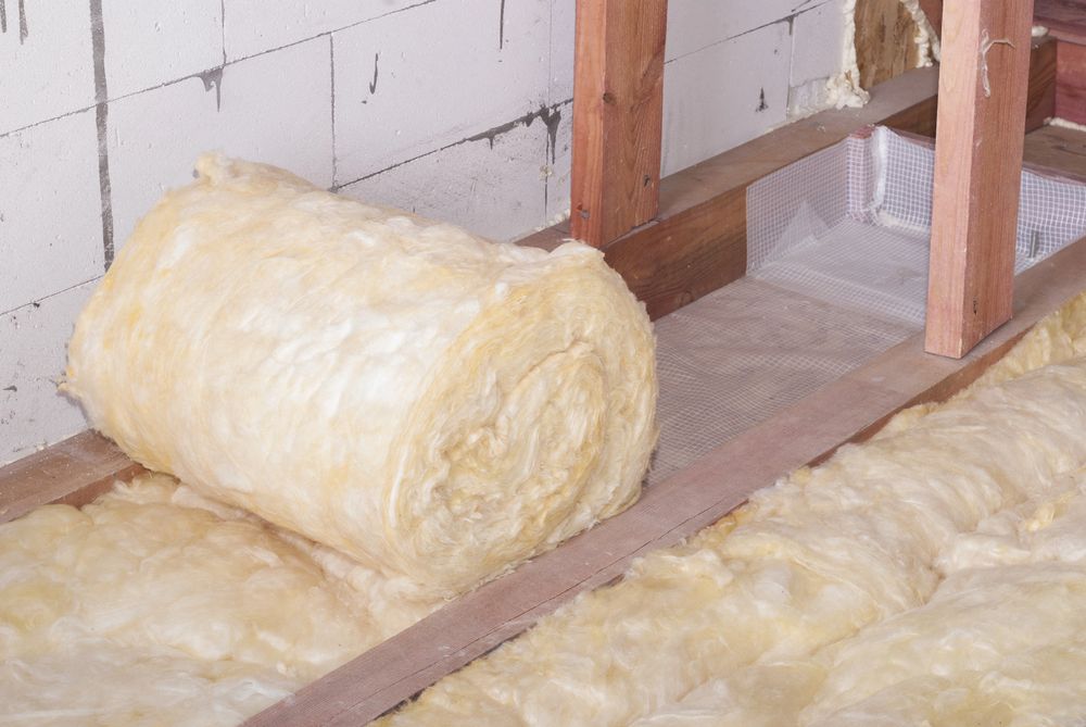 A roll of fiberglass insulation, partially unrolled by an insulation contractor, rests on the floor between wooden studs and a concrete wall in a building under construction.