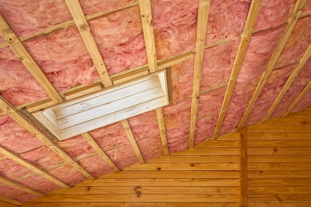 Exposed ceiling with pink fiberglass insulation installed between wooden beams by an insulation contractor, featuring a skylight frame and wood-paneled walls below.
