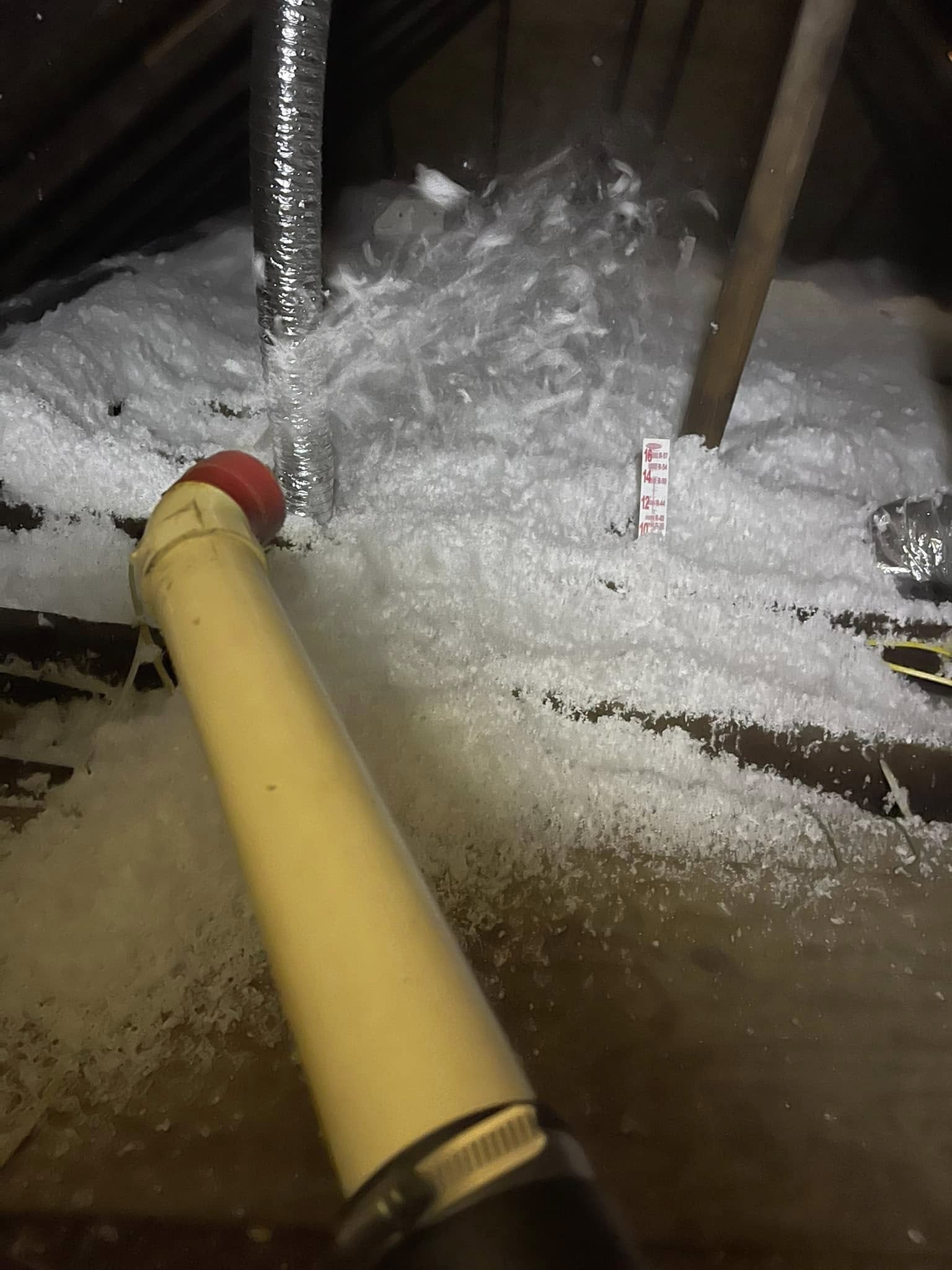 A yellow insulation blower tube lays in an attic, blowing white loose-fill insulation near a depth gauge, with ductwork visible in the background.