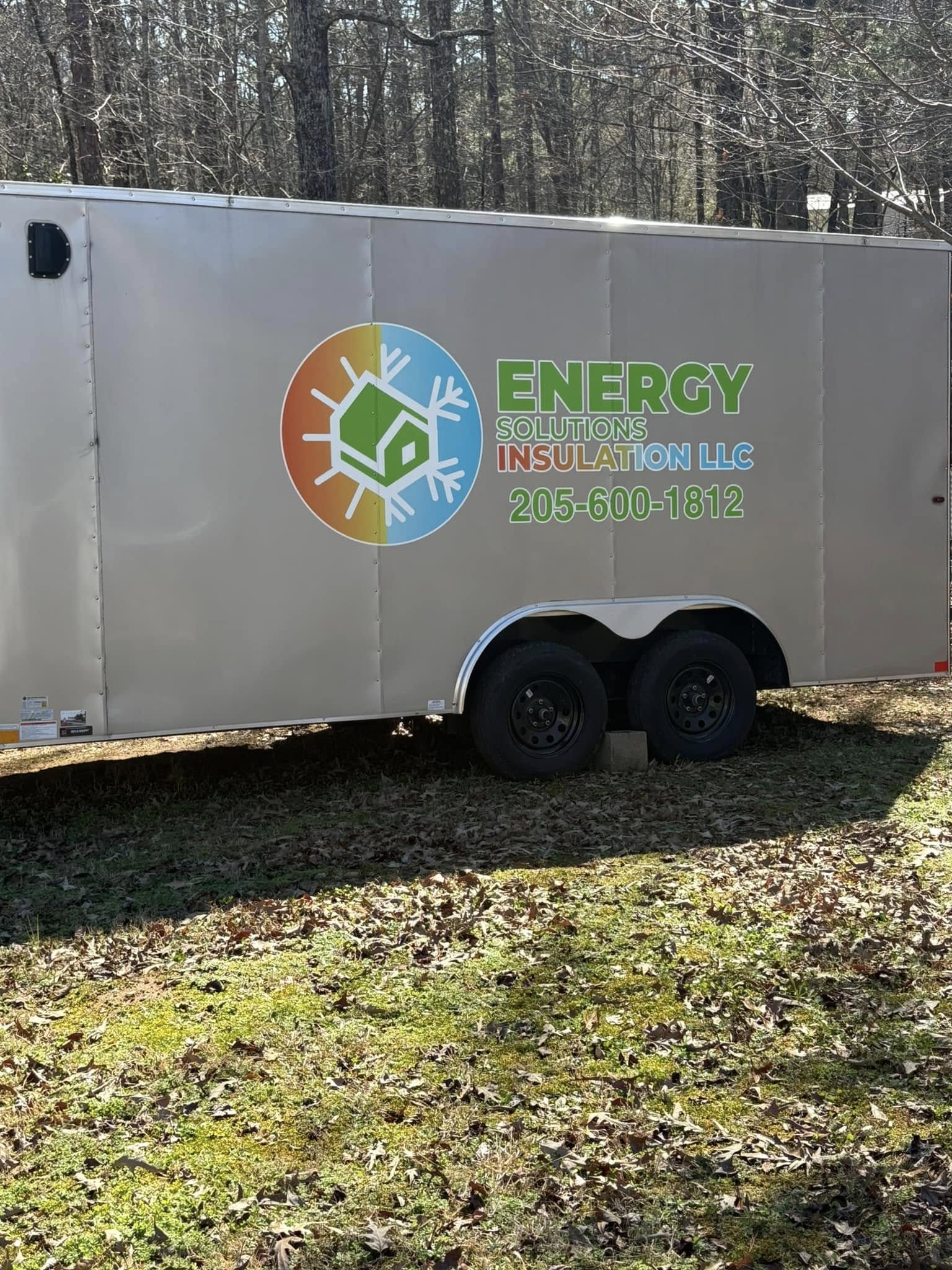 A beige trailer with Energy Solutions Insulation LLC logo and contact number 205-600-1812 parked on grass with trees in the background